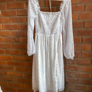 Vit blommig långklänning Vero Moda - Supersöt långklänning från Vero Moda i vitt med ljusblått blommigt mönster. Klänningen har smockad överdel, långa puffärmar och en liten slits framtill. Perfekt för sommarens alla tillfällen, med luftig och skön känsla.