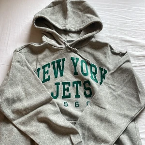 Grå New York Jets hoodie med tryck - Snygg grå hoodie med New York Jets-tryck i grönt framtill och årtalet 1960. Tröjan har huva med snörning och en stor magficka. Perfekt för dig som gillar sportig och avslappnad stil.