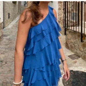 Blå oneshoulder volangklänning - Säljer en blå kortklänning med flera lager volanger och oneshoulder-design. Klänningen har en loose passform och är perfekt för dig som gillar att sticka ut med en cool och trendig look. Materialet känns lätt och luftigt, vilket gör den skön att bära under varma dagar.