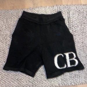 Svarta stickade shorts CB - Svarta stickade shorts i medium med vit CB-logga framtill. Modellen har två bakfickor och elastisk midja för skön passform. Materialet är mjukt och stickat, perfekt för chill dagar. Snygg och enkel design med cool branding.