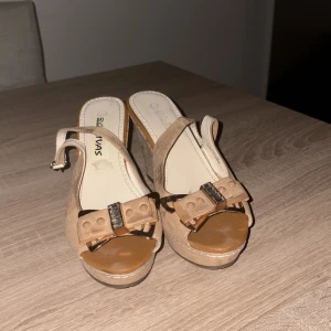 Beige sandaler med kilklack och rosett - Snygga beige sandaler med kilklack i korklook och öppen tå. Skorna har en dekorativ rosett framtill med glittriga detaljer och justerbar rem runt hälen. Ovandelen är i mocka och innersulan är ljus med logga. Perfekta för sommarens alla tillfällen.