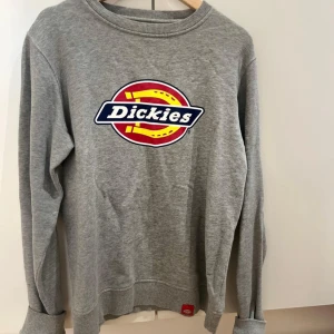 Grå Dickies sweatshirt i bomull - Säljer en grå sweatshirt från Dickies i 100% bomull. Tröjan har klassisk Dickies-logga i rött, gult och blått framtill, rund hals och långa ärmar. Perfekt för dig som gillar streetwear och enkel stil.