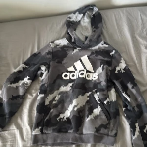 Camouflage hoodie från Adidas - Säljer en grå, svart och vit hoodie från Adidas med stort logotryck på bröstet. Tröjan har ett fett camouflage-mönster, huva och magficka. Perfekt för dig som gillar streetwear och sportig stil. Materialet är mjuk bomull och polyesterblandning.
