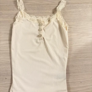 Beige ribbad linnetopp med spets - Supersöt beige linnetopp med tunna axelband och spetsdetaljer längs urringningen. Ribbad struktur och tre små knappar framtill ger en extra touch. Perfekt att styla lager på lager eller som den är under varma dagar.