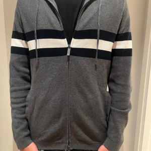 Grå hoodie med ränder och dragkedja från Massimo Dutti - Grå hoodie med svarta och vita horisontella ränder över bröstet. Tröjan har huva med snörning, hel dragkedja framtill och långa ärmar. Perfekt för en chill och avslappnad stil. Nästan som ny, aldrig använd. 