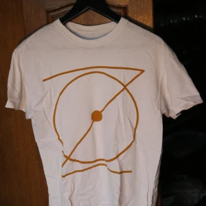 Vit t-shirt med grafiskt tryck från band - Vit t-shirt med korta ärmar och ett stort grafiskt tryck i senapsgult på framsidan. Trycket består av cirklar och linjer som ger en modern och stilren vibe. T-shirten har klassisk passform och är gjord i mjuk bomull, perfekt för en chill look. Lappen är bortklippt.