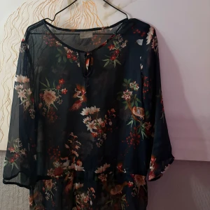 Blommig mörkblå blus Vero Moda - Snygg mörkblå blus från Vero Moda med blommigt mönster och fåglar. Blusen  är i tunn, transparent polyester med knytning vid halsen och trekvartsärmar. Perfekt för dig som gillar en luftig och trendig stil med fina detaljer.