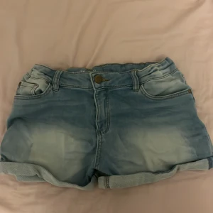 Blå jeansshorts från Lindex - Snygga blå jeansshorts från Lindex med klassisk femficksdesign och uppvikta benslut. Sjortsen har som ett slit märke på vänstra sida och högra sida även ett i fram fickan synns på första bilden!!Shortsen har normal passform men kan nog även passa nån storlek större och är tillverkade i mjukt denimtyg. Perfekta för varma dagar och enkla att matcha med allt.
