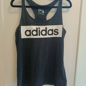 Svart linnetopp från Adidas - Sportig svart linnetopp från Adidas med stort vitt logotryck på bröstet. Modellen är ärmlös med brottarrygg och tillverkad i mjuk bomull som andas. Perfekt för gymmet eller andra aktiva dagar.