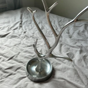 Smyckesträd i silverfärgad metall - Snyggt smyckesträd i silverfärgad metall med grenar för att hänga ringar, armband, halsband och örhängen. Modern och stilren design som liknar ett träd, perfekt för att hålla koll på dina favoritsmycken. Rund fot som står stadigt på bordet.
