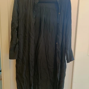 Svart långklänning med knappar - En svart långklänning med långa ärmar och knäppning hela vägen framtill. Klänningen har en enkel och stilren design med slits vid ärmslutet. Materialet känns lätt och har en diskret struktur, perfekt för dig som gillar minimalistisk stil.