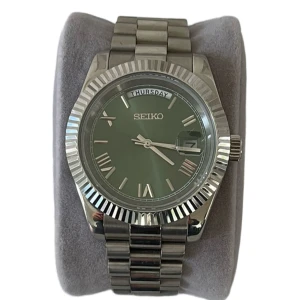 Oliv-Grön Seiko Mod Day-Date - Stilren Seiko klocka med oliv-grön urtavla, romerska siffror och dag/datumvisning. Inspererad av Day-Date. Boetten och länken är i blankt silverfärgat stål och urtavlan har en snygg solreflex. Perfekt accessoar för dig som vill sticka ut med en klassisk men modern look. Alla länkar + box tillkommer självklart vid köp‼️🙌 
