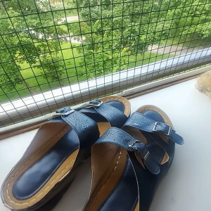 Mörkblå sandaler med spännen - Snygga mörkblå sandaler med två breda remmar i skinn och justerbara metallspännen. Remmarna har dekorativa hålmönster och sulan är beige med synliga sömmar. Perfekta för varma dagar och ger en stilren look.