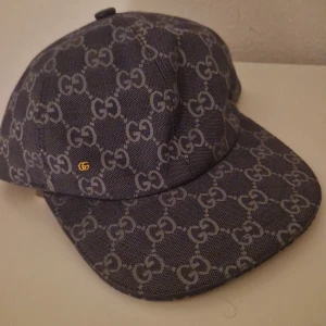 Gucci keps med GG-mönster - Snygg keps från Gucci i mörkblått tyg med ikoniskt GG-mönster över hela kepsen. Klassisk form med böjd skärm och liten guldfärgad GG-detalj på sidan. Tillverkad i bomull och polyester för en exklusiv känsla.