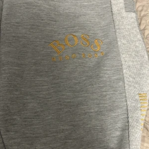 Grå mjukisbyxor från Hugo Boss - Snygga grå mjukisbyxor från Hugo Boss med guldfärgad logga på benet och detaljer längs sidorna. Byxorna har elastisk midja, muddar vid bensluten och sidofickor. Perfekta för en chill och stilren look. Pris kan diskuteras.