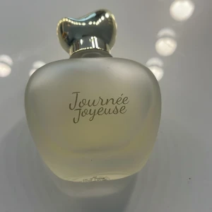 Journée Joyeuse Eau de Parfum - Elegant parfym i frostat glas med mjuka former och en unik, silvrig kork. Flaskan har en lyxig känsla och texten 'Journée Joyeuse' på framsidan. Perfekt för dig som gillar stilrena och moderna doftflaskor.