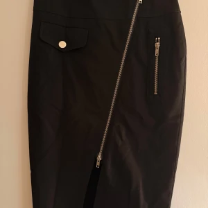 Svart kjol Jean Paul Gaultier x Lindex - Svart kortkjol från Jean Paul Gaultier x Lindex med cool asymmetrisk dragkedja, fickdetaljer och bälte i midjan. Kjolen har en edgy vibe med silvriga metalldetaljer och passar perfekt för dig som gillar statement-plagg. (Köpt ny) Aldrig använd