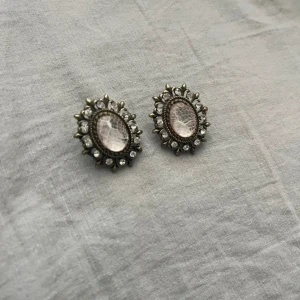 Vintage-inspirerade ovala örhängen - Säljer ett par ovala örhängen med antikguld-färgad metall och glittriga vita stenar runt en ljusrosa, mönstrad mittdel. De har en elegant, retro vibe och är perfekta för att lyfta din outfit med lite extra bling.