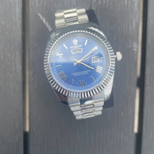  Binbond klocka - Stilren klocka från Binbond med blå urtavla, romerska siffror och dag- och datumvisning. Boetten och länken är i silverfärgat stål med räfflad kant runt urtavlan. Klassisk design som passar till många olika outfits.