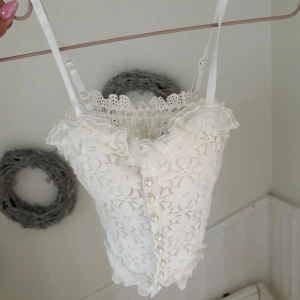 Vit spetsig mjuk korsett med pärlknappar - Supersöt vit korsettopp med blommig spets, volangkant upptill och pärlknappar framtill. Smala axelband och romantisk känsla, perfekt för dig som gillar detaljer och vintage vibes.