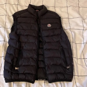 Svart dunväst från Moncler - Snygg svart dunväst från Moncler med hög krage och klassisk logga på bröstet. Västen har två dragkedjefickor framtill, quiltad design och diskreta Moncler-detaljer längs ärmöppningarna. Perfekt att slänga över en hoodie eller tröja för extra värme.