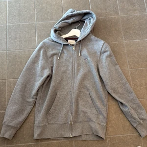 Grå hoodie från GANT med dragkedja - Säljer en stilren grå hoodie från GANT med huva och snörning. Tröjan har dragkedja framtill, två fickor och ett litet broderat GANT-emblem på bröstet. Perfekt för chill dagar och enkel att matcha med jeans eller joggers. Kvitto finns från boozt. Nypris i butik: 1600-800. Storlek S