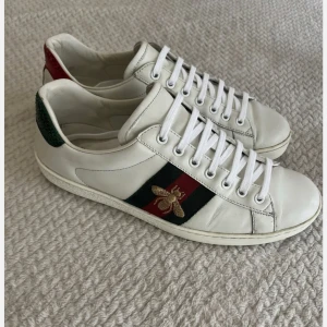 Gucci Ace sneakers med bi-detalj - Säljer ett par Gucci Ace sneakers i vitt skinn med klassiska gröna och röda ränder på sidan och en broderad guldfärgad bi. Skorna har snörning och platt sula, samt gröna och röda detaljer på hälen. Perfekt för dig som gillar lyxiga och trendiga sneakers.