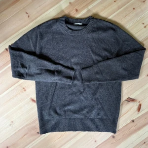 Mörkgrå stickad tröja i ull - En stilren mörkgrå stickad tröja med rund hals och ribbade muddar vid ärmslut och nederkant. Perfekt för lager-på-lager och passar till både jeans och chinos. Tröjan är gjord i mjuk ull och har en klassisk, avslappnad passform.