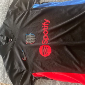FC Barcelona svart Nike matchtröja - Snygg FC Barcelona fotbollströja i svart med röda och blå detaljer. Tröjan har korta ärmar, klubbmärke på bröstet och röd Spotify-logga framtill. Tillverkad i lätt och ventilerande material, perfekt för träning eller match. Klassisk Nike Swoosh på bröstet.