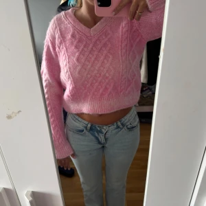 Rosa croppad stickad tröja MNG - Superfin rosa stickad tröja från MNG med v-ringning och kabelstickat mönster. Croppad modell med långa ärmar och ribbade muddar. Gjord i återvunnen polyester, perfekt för dig som gillar en söt och trendig look.