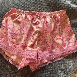Rosa pyjamasshorts med spets - Superfina shorts i satin material i rosa, gult och orange. De har söt rosa rosett med hjärtberlock i midjan och bred spetskant nertill. Perfekta för sommarkvällar eller som pyjamasshorts.