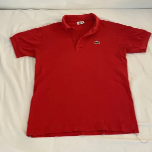 Röd pikétröja från Lacoste - Klassisk röd pikétröja från Lacoste med broderad krokodil på bröstet. Tröjan har korta ärmar, krage och knäppning framtill. Tillverkad i bomull för en skön och luftig känsla. Perfekt för dig som gillar stilren och sportig look.