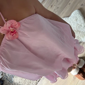 Rosa volangklänning med blomdetalj - Supersöt kort rosa klänning med volanger och halterneck. Klänningen har en stor dekorativ blomma vid halsen och är tillverkad i ett lätt, transparent material som ger en fluffig och luftig känsla. Perfekt för dig som vill sticka ut med en lekfull och feminin look.