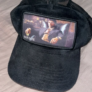 Svart keps från Rovior med motiv - Svart keps från Rovior med mesh baktill och framsida i manchester. Stor patch framtill med motiv av en man i kostym och texten Rovior. Snygg detalj med ett silverfärgat R på sidan. Perfekt för dig som vill sticka ut.