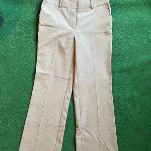 Beige kostymbyxor från KappAhl - Stilrena beige kostymbyxor från KappAhl i rak modell med pressveck och hällor för bälte. Byxorna har sidfickor och är tillverkade i ett mjukt tyg som ger en dressad look. Perfekta för dig som gillar klassisk och clean stil.