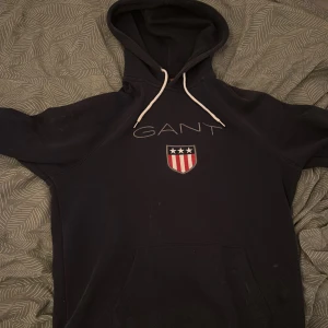 Svart hoodie från GANT med emblem - Svart hoodie från GANT med vit snörning och stort broderat emblem på bröstet. Tröjan har huva, känguruficka och GANT-logga med amerikanskt inspirerat sköldmärke i rött, vitt och blått. Perfekt för chill dagar och streetstyle.