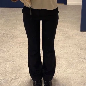 Svarta kostymbyxor med bootcut - Snygga svarta kostymbyxor i modellen Vera med bootcut-snitt och klassisk midja. Byxorna har bälteshällor och en stilren look som passar till många olika outfits. Perfekta för dig som gillar en clean och tidlös stil. Storlek 34, Inga defekter!