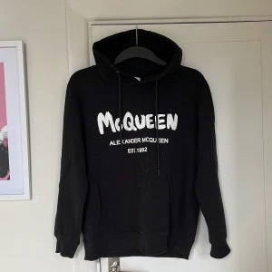 Svart hoodie Alexander McQueen - Svart hoodie från Alexander McQueen med vit logga framtill. Klassisk modell med huva, snörning och stor magficka. Långärmad och ribbade muddar. Perfekt för dig som gillar streetstyle och vill ha en snygg statementtröja. Passar XS och mindre, hör av er vid frågor