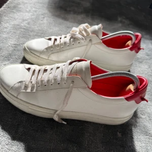 Vita Givenchy sneakers  - Snygga vita sneakers från Givenchy med röd häl och guldfärgad logga på baksidan. Skorna har klassisk rund tå, platt sula och snörning framtill. Tillverkade i skinn med stilren design och lyxig känsla. Perfekta för dig som gillar exklusiva och trendiga sneakers.