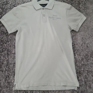 Ljusgrå pikétröja Peak Performance - Snygg ljusgrå pikétröja från Peak Performance med klassisk krage och två knappar framtill. Broderad logga på bröstet. Tillverkad i mjuk bomull, perfekt för en clean och stilren look. Kortärmad modell som passar året runt.