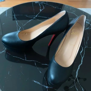 Svarta pumps  - Klassiska svarta pumps ikonisk röd sula. Skorna har rund tå, hög klack och en stilren, elegant design som lyfter varje outfit. Perfekta för dig som vill ha en tidlös och lyxig look.