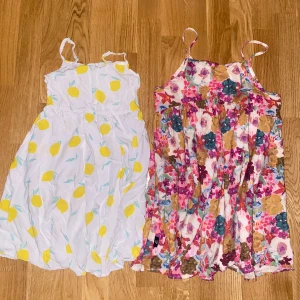 Två somriga kortklänningar med mönster - Säljer två söta kortklänningar med smala axelband. Ena klänningen är vit med ett fräscht citrongult mönster, den andra har ett färgglatt blommigt mönster i rosa, blått, lila och brunt. Båda är perfekta för varma dagar och har en luftig passform. 5/6 år