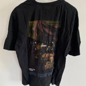 Svart t-shirt Gallery Dept Arise - Svart t-shirt från Gallery Dept med tryck både fram och bak. Bakstycket har ett stort konstnärligt motiv. Framsidan har ett mindre tryck med grafiska element. Klassisk rund hals och korta ärmar. Perfekt för dig som gillar streetwear och unika prints.