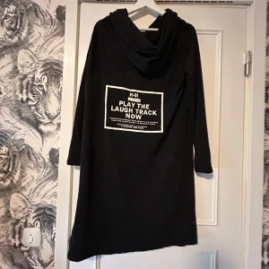 Svart lång Cape/tröja/cardigan med stor huva och tryck - Svart lång cape/tröja/cardigan med stor huva och långa ärmar. På ryggen finns ett stort vitt tryck med texten 'PLAY THE LAUGH TRACK NOW'. Plagget har en avslappnad passform och är perfekt för dig som gillar streetstyle och statement-plagg. 