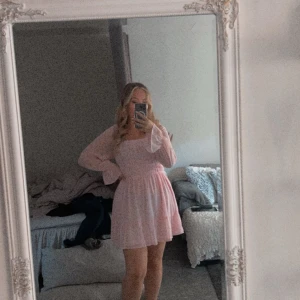 Rosa kort klänning från Eillybazar - Supersöt ljusrosa klänning från Eillybazar med smockad överdel och volangdetaljer. Klänningen har långa, transparenta ärmar med lätt volang vid ärmslut och en flowy kjol. Perfekt för dig som gillar romantisk och feminin stil. Storlek L men också väldigt strechig. Använd en gång 