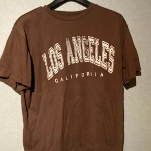 Brun Los Angeles t-shirt i bomull - Snygg brun t-shirt med trycket 'Los Angeles California' i vitt på framsidan. Klassisk rund hals och korta ärmar. Tillverkad i mjuk bomull som är skön mot huden. Perfekt för dig som gillar streetstyle och avslappnad vibe.
