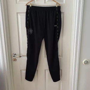 Svarta träningsbyxor Hammarby - Svarta mjukisbyxor från Craft med elastisk midja och logga på benet. Snygga detaljer med logoband längs sidorna och en sportig look. Perfekta för träning eller chill, tillverkade i mjukt och lätt material som andas.