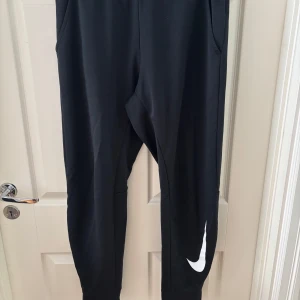 Svarta mjukisbyxor från Nike - Svarta mjukisbyxor från Nike med klassisk vit swoosh-logga på ena benet. Byxorna har elastisk midja, smal passform och är tillverkade i mjukt material som passar perfekt för chill eller träning. Snygg och enkel design med fickor på sidorna.