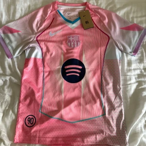 Rosa Barça Ställ (#10 Lamine Yamal) - Snygg FC Barcelona fotbollströja från Nike i rosa med vita och ljusblå detaljer. Kortärmad med V-ringad krage och tryckta ränder. Spotify-logga på bröstet och Lamine Yamal 10 på ryggen. Tillverkad i lätt, ventilerande polyester. Perfekt för dig som älskar Barça.