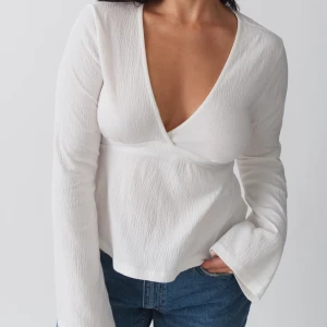 Vit Tie Topp från Gina Tricot - Supersnygg vit omlottopp från Gina Tricot med lång ärm och djup v-ringning. Toppen har knytband i midjan och är gjord i ett lätt, krinklat material som ger en cool textur. 